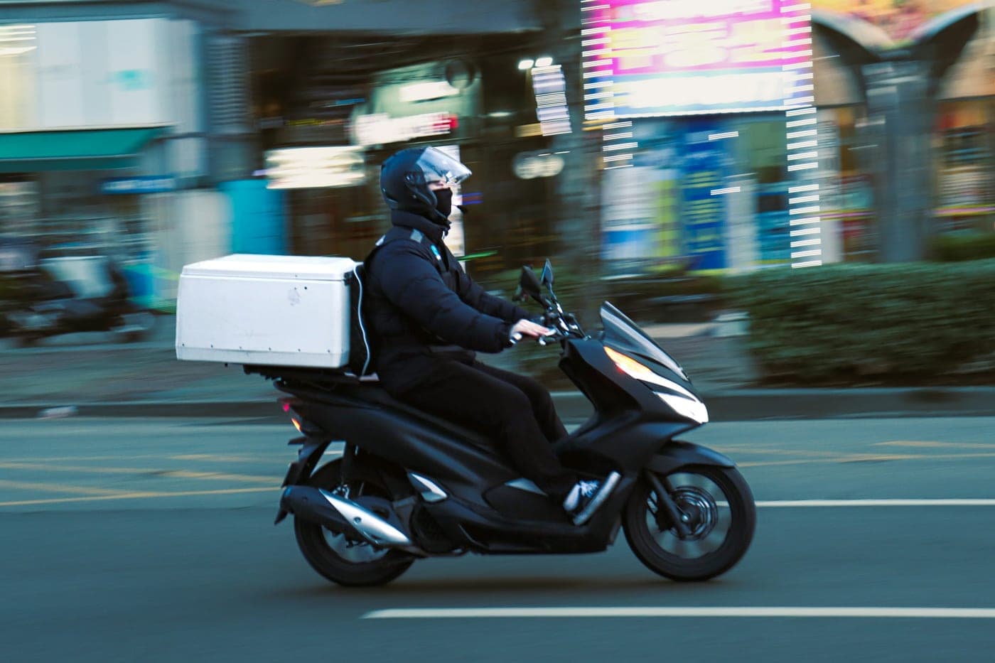 Entregador de scooter preta com baú branco circulando por uma avenida