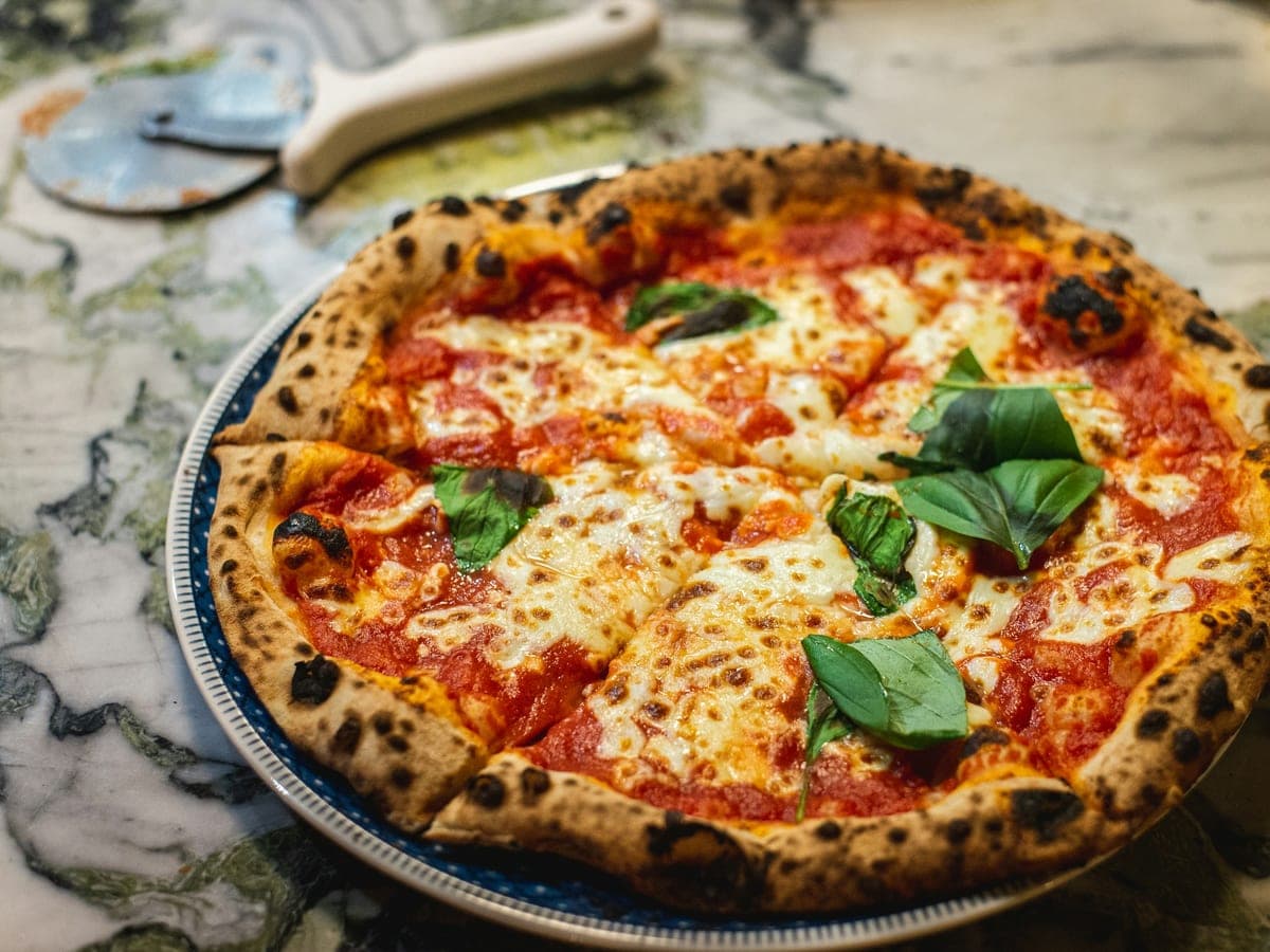 Pizza margherita com manjericão fresco em prato azul, com cortador de pizza ao fundo